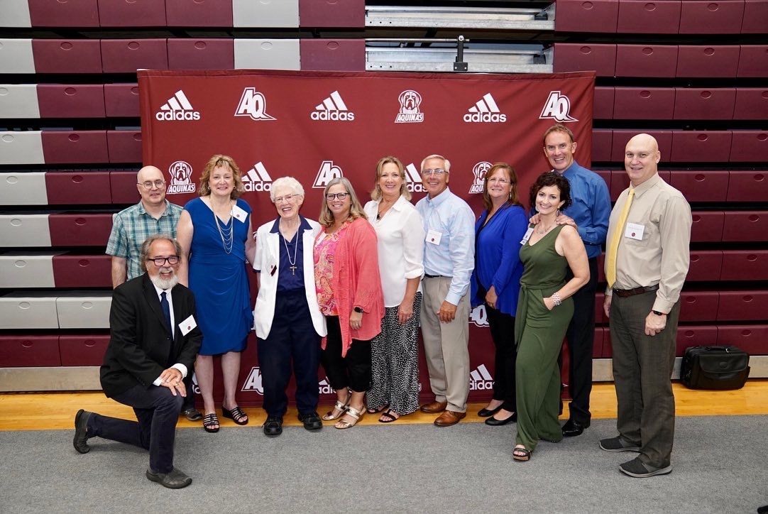 A group of smiling people, including Sister Ann Mason, posing for a photo in front of a Red AQ/Adidas printed backdro