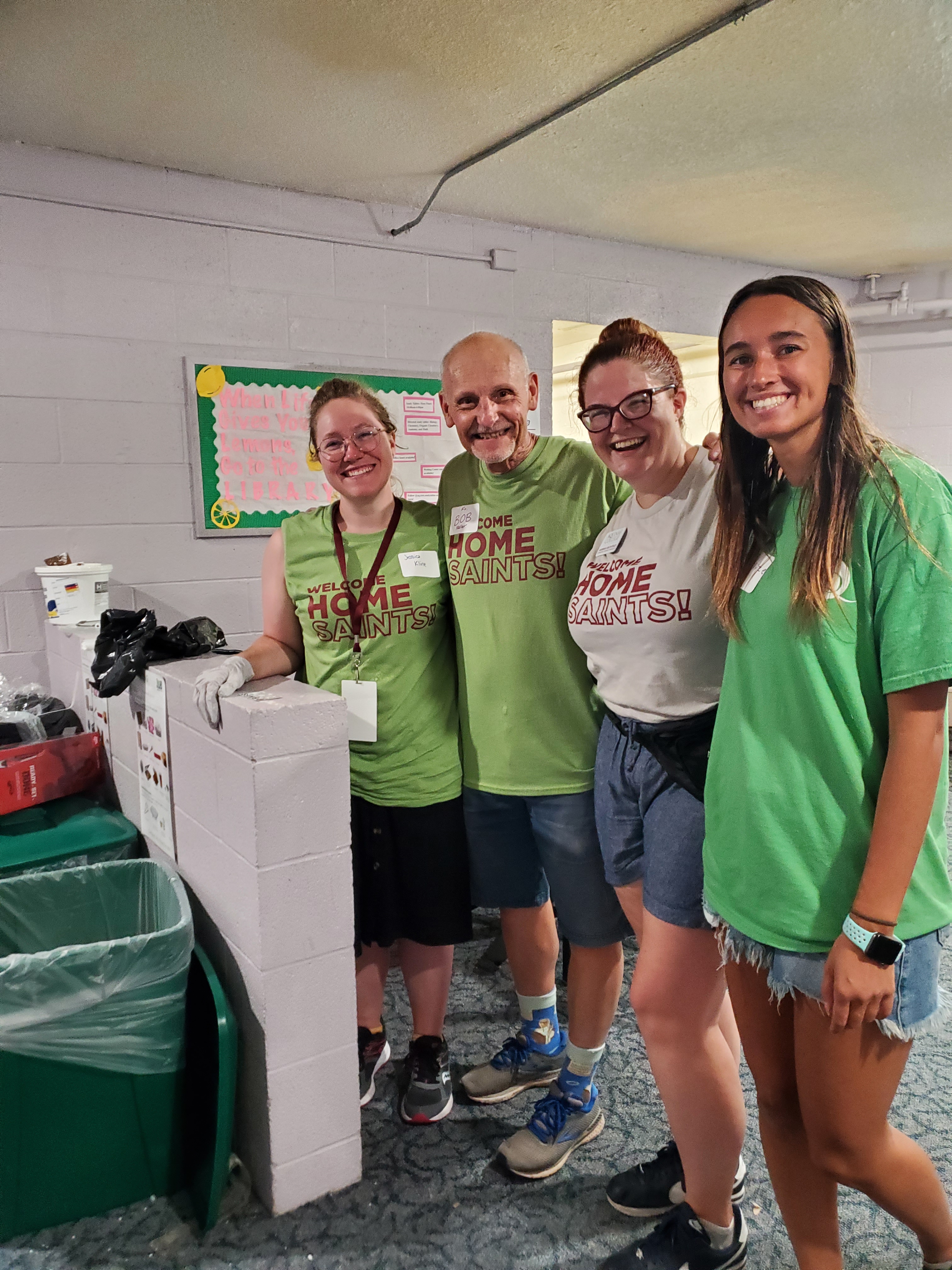 Four people smiling in shorts, sneakers, AQ T shirts next to recycling bins