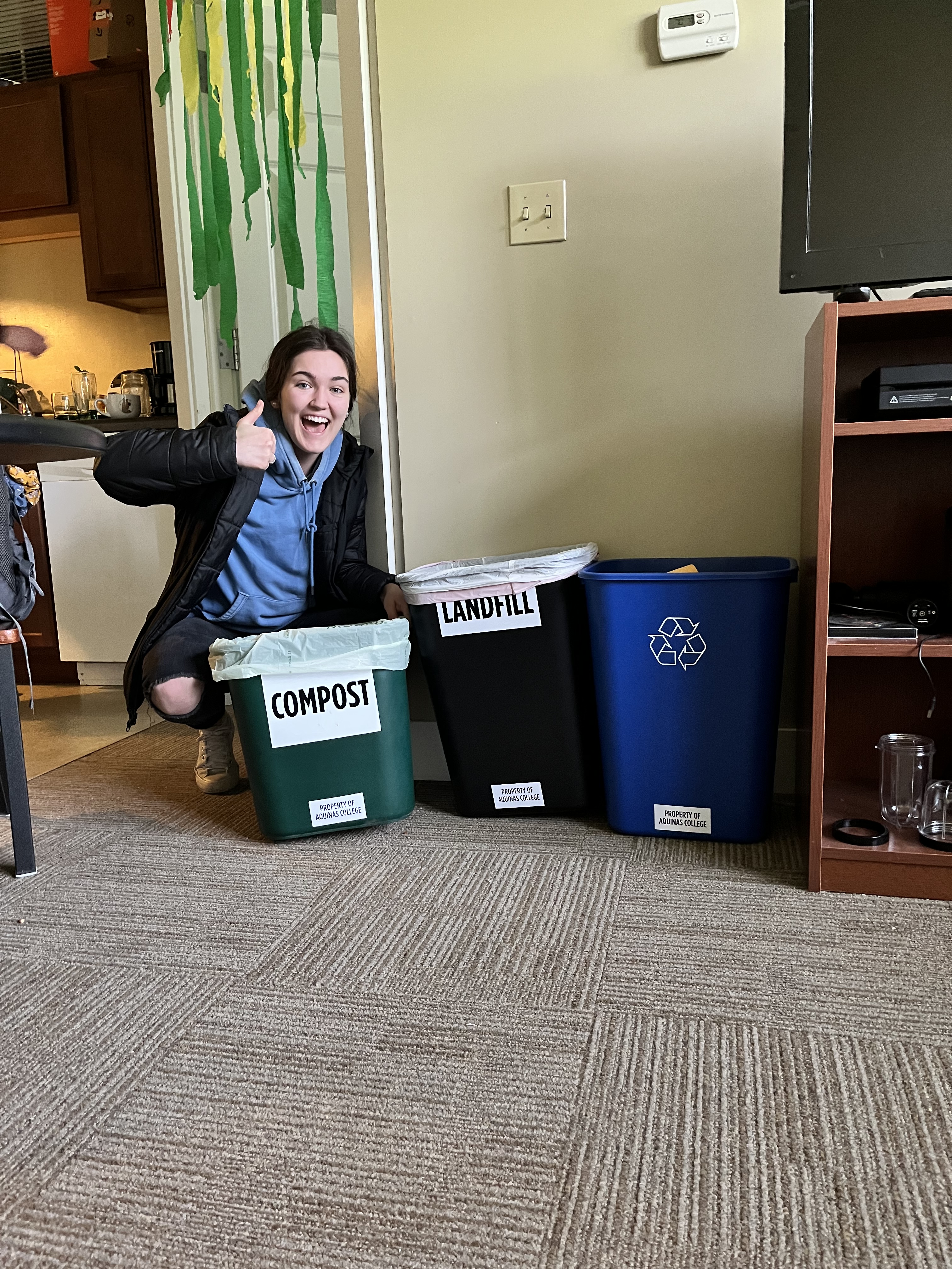 Student giving a thumbs up and sitting on the floor of their home or dorm with three bins labeled compost, landfill, and recycling. 