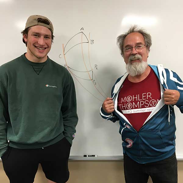 A smiling student and professor stand in front of a whiteboard with a diagram written on it. The professor has unzipped his hoodie to reveal his Mohler Thompson AQ summer Research T Shirt. 