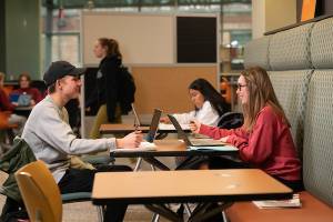 Two students sitting across from one another at a table smiling with their laptops open