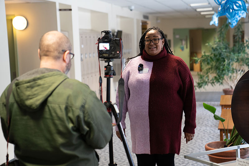 Alicia Lloyd speaking in front of a camera in a hallway while someone operates the camera