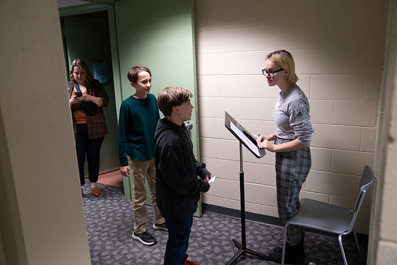 Student at a music stand listening to child talking holding a paper card. He stands next to another child. A woman looks at her phone in the doorway.