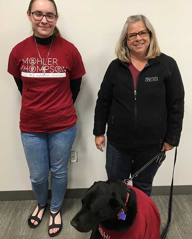 A student and a professor stand side by side in Aquinas shirts against a blank wall. The professor holds a leash for a black lab wearing an Aquinas Red T shirt. 