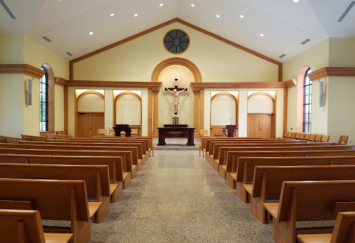A view down the aisle of Our Lady Seat of Wisdom Chapel. Warm wood pews on either side. Christ on the Cross looms over the alter. The walls are a yellowish beige lined with warm wood details.