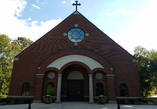 Simple brick chapel with a circular window above an arched doorway. A cross stands at the peak of the roof. Blue skies and white clouds behind.