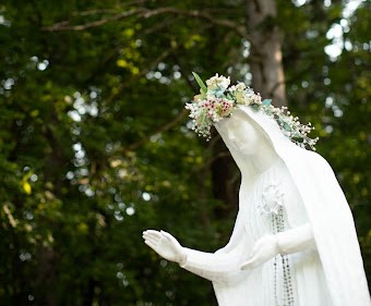 Our Lady of Fatima Statue adorned with beads and a flower crown with a backdrop of green tree foliage.