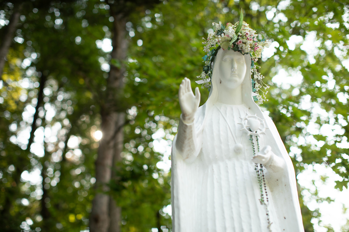 Statue of our lady fatima with a crown of flowers and rosary and a backdrop of leaves