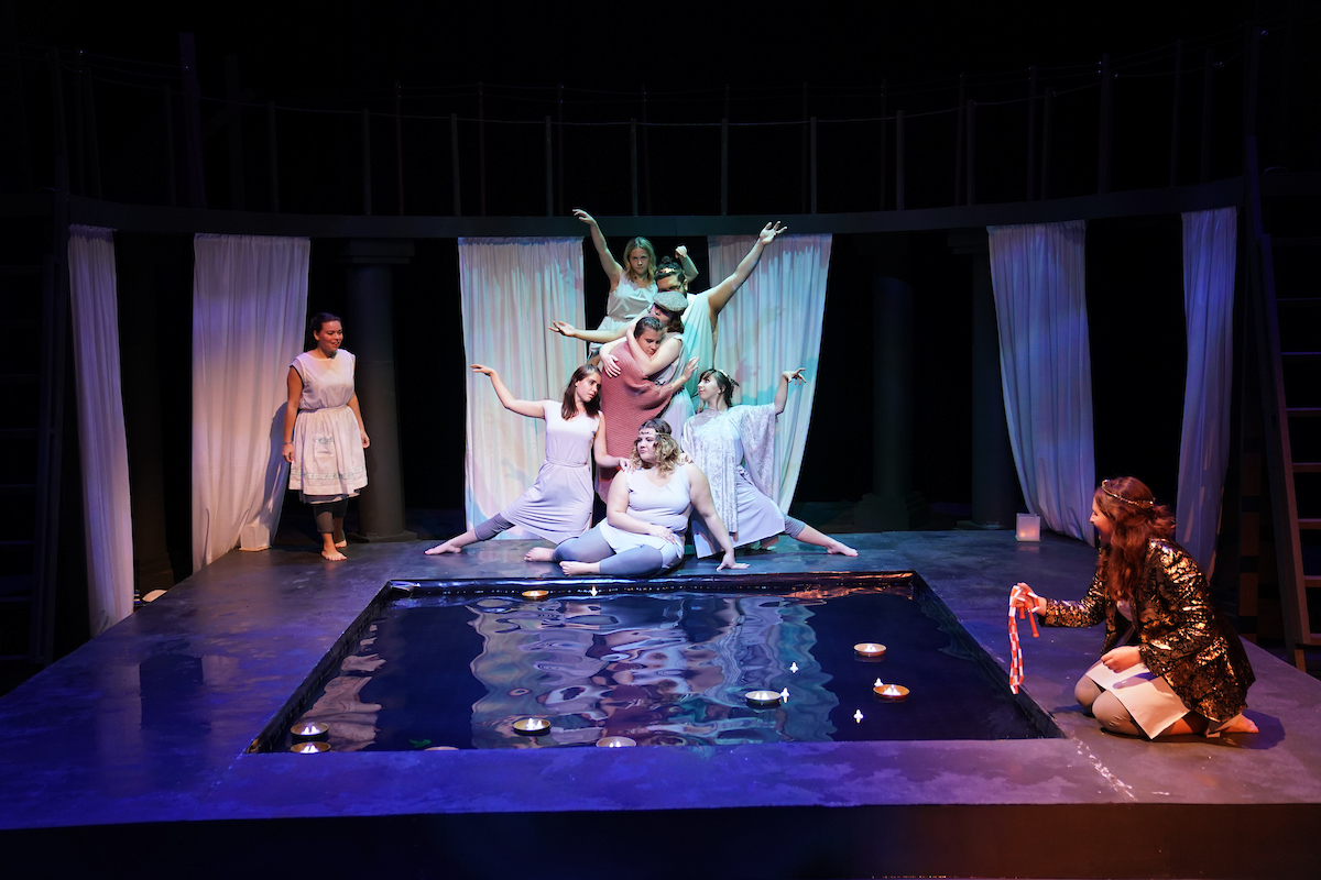 Actors in clothes that look like white togas pose dramatically at the end of a pool of water.
