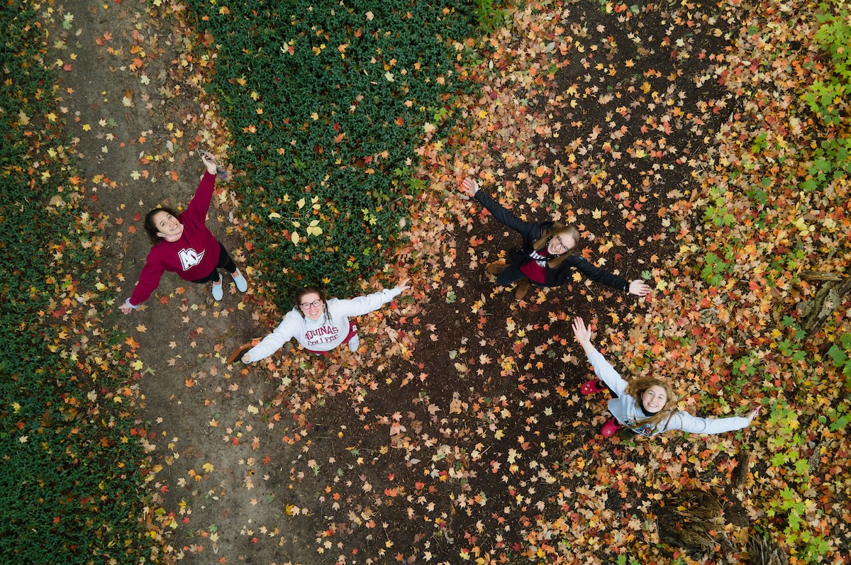 Overhead shot of three students with arms outstretched on a leafy trail in fall smiling up at the camera