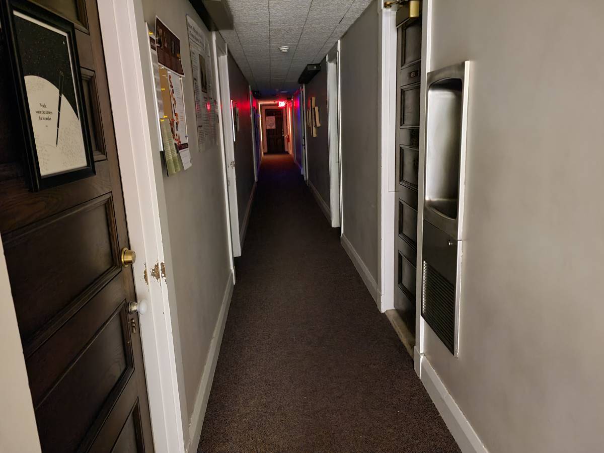 Photo of the 3rd floor hallway at night. Narrow and full of doors. A glowing red exit sign at the end.