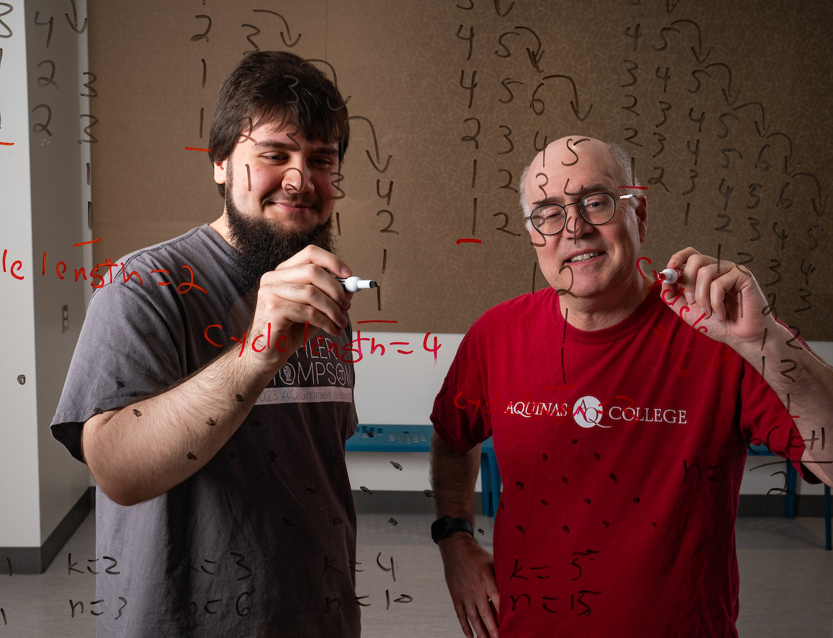 Mehanovic and Spencer holding dry erase markers up to a clear glass board full of numbers