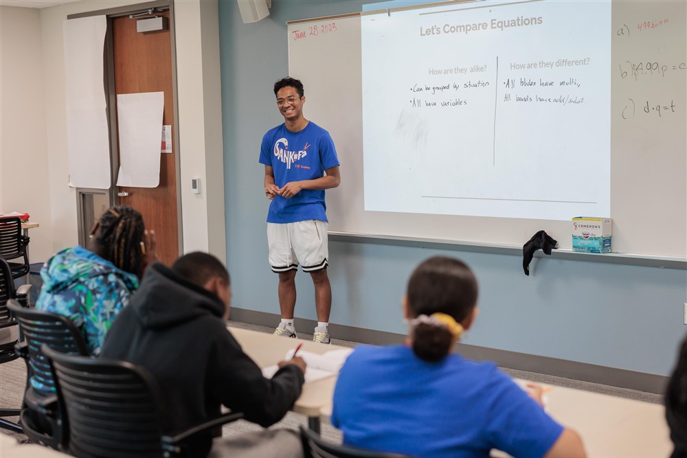 Students listening to a teacher who is smiling in front of a whiteboard