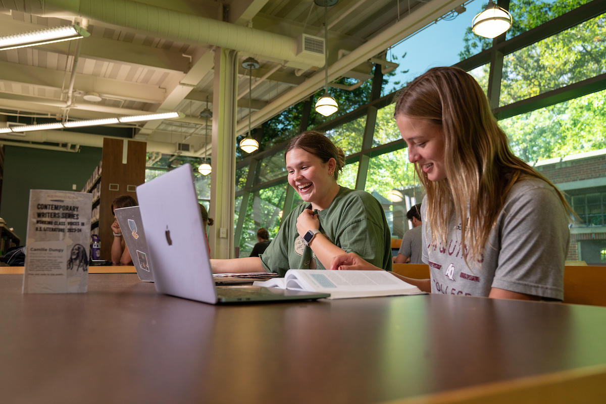 Two students talk at a table in the library with laptops