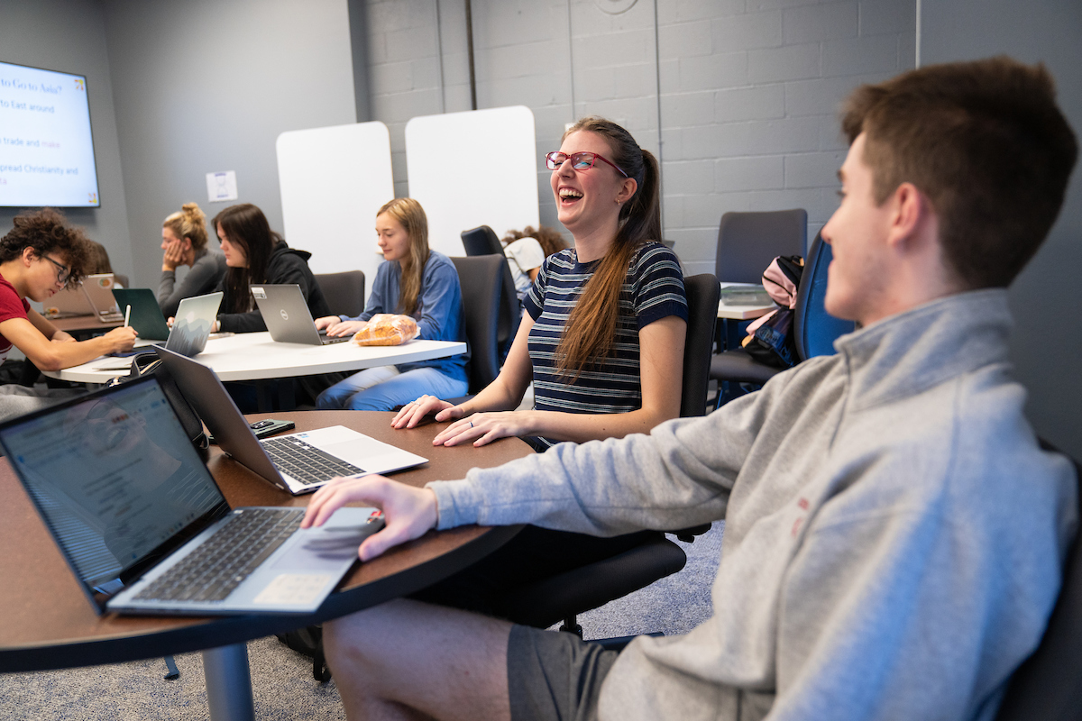 Student smiling and laughing at desk with laptop. More students in the background and foreground.